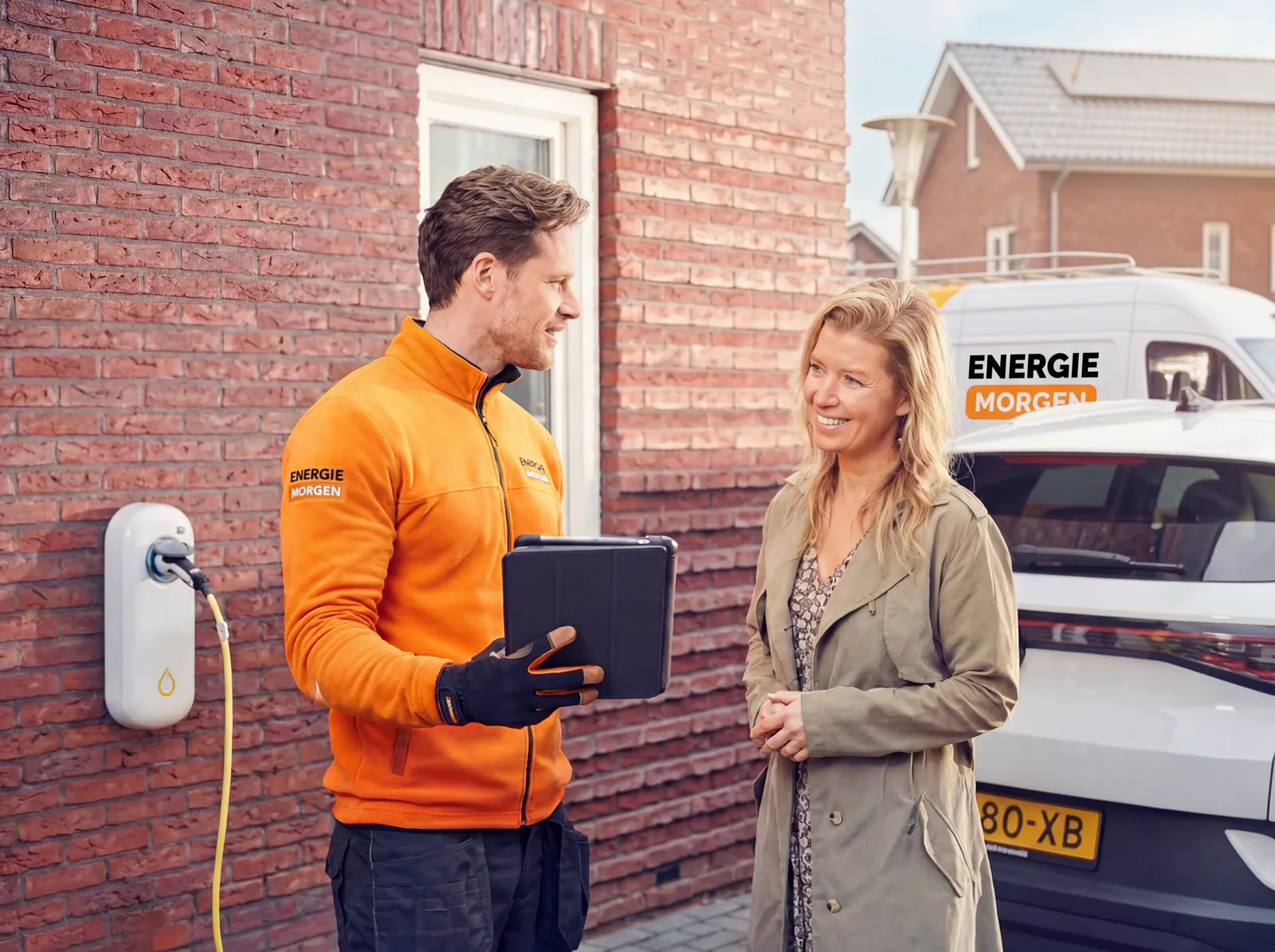 Monteur van Energie Morgen in bedrijfskleding met tablet bij een klant thuis, met de servicebus op de achtergrond