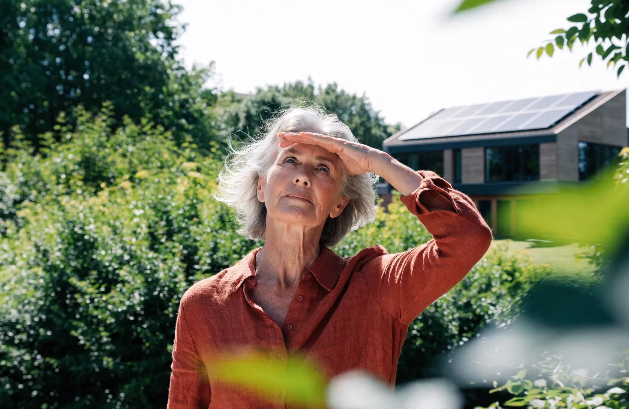 Vrouw kijkt met de hand boven haar ogen naar de zon, bij een woning met zonnepanelen