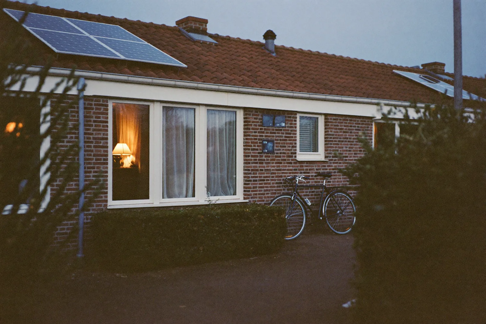 Nederlandse rijtjeswoning in de schemering met zonnepanelen op het dak en warm lamplicht achter het raam